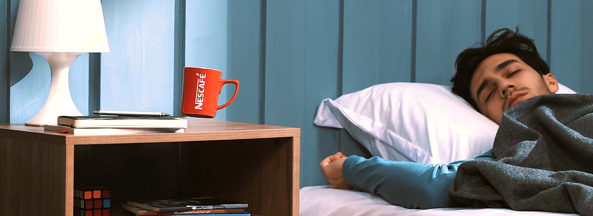 A boy asleep in his bed with a red nescafe mug in mid-air on the bedside table